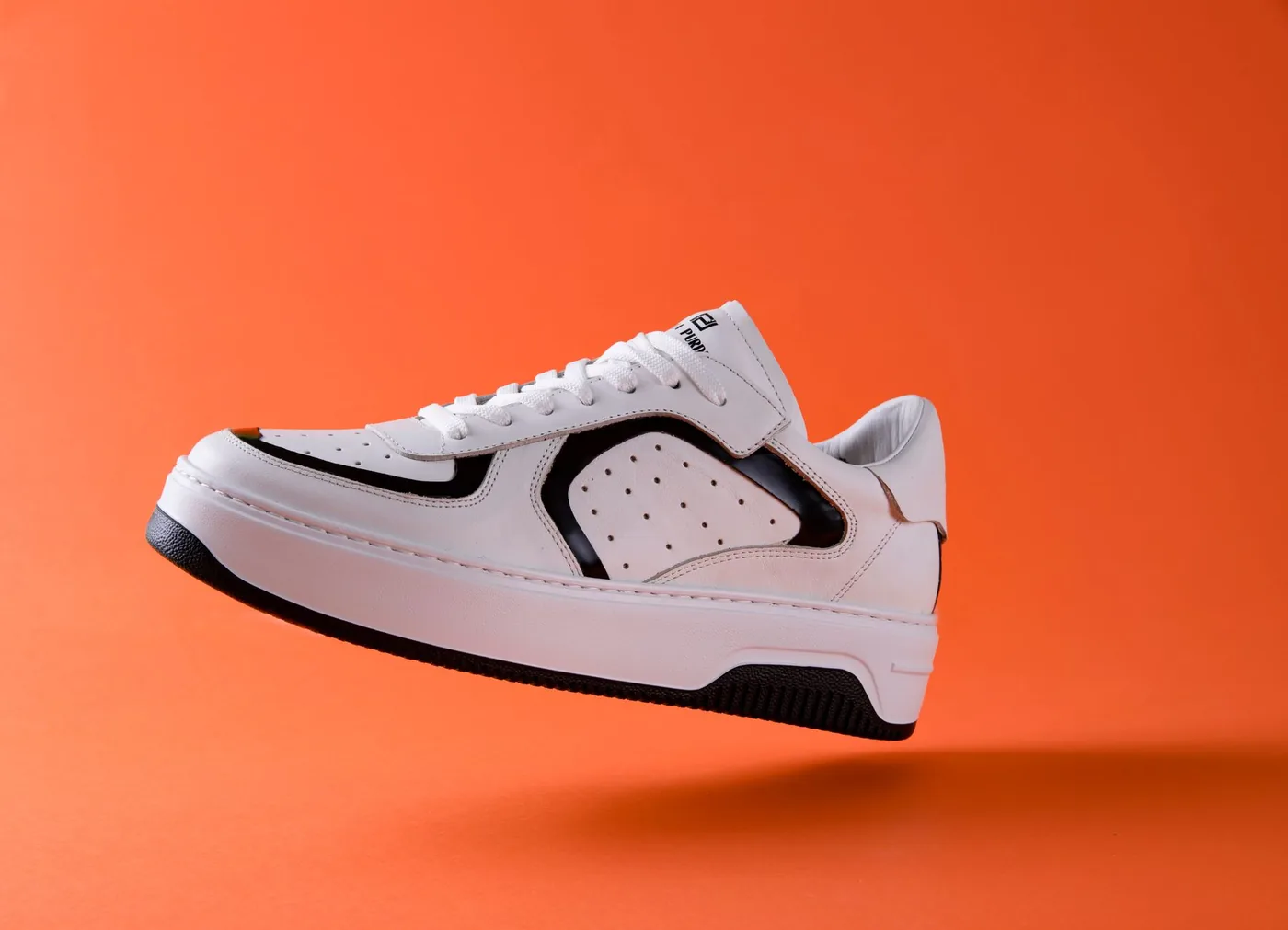 Zapatilla blanca con detalles negros flotando sobre fondo naranja, ejemplo de producto que se vende por detalle